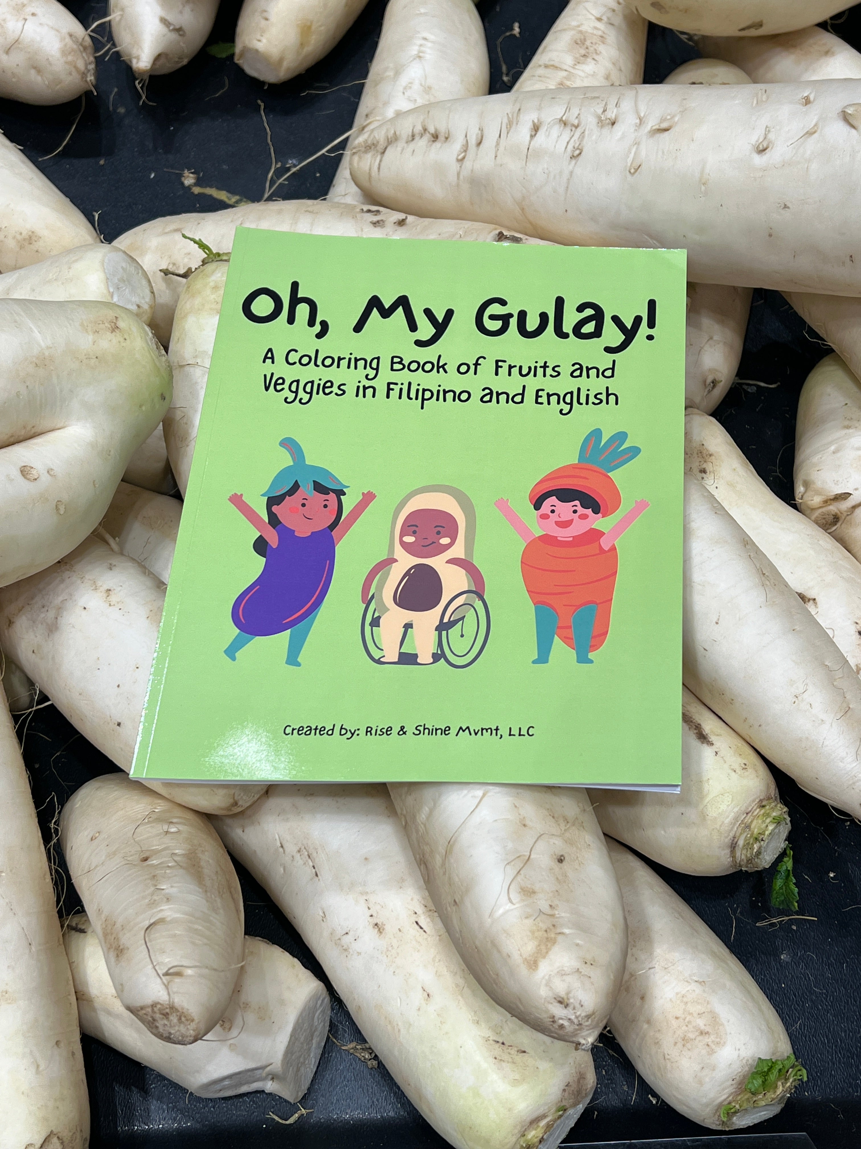 A copy of the green cover "Oh, My Gulay" coloring book is displayed on top of white daikon radishes at a supermarket